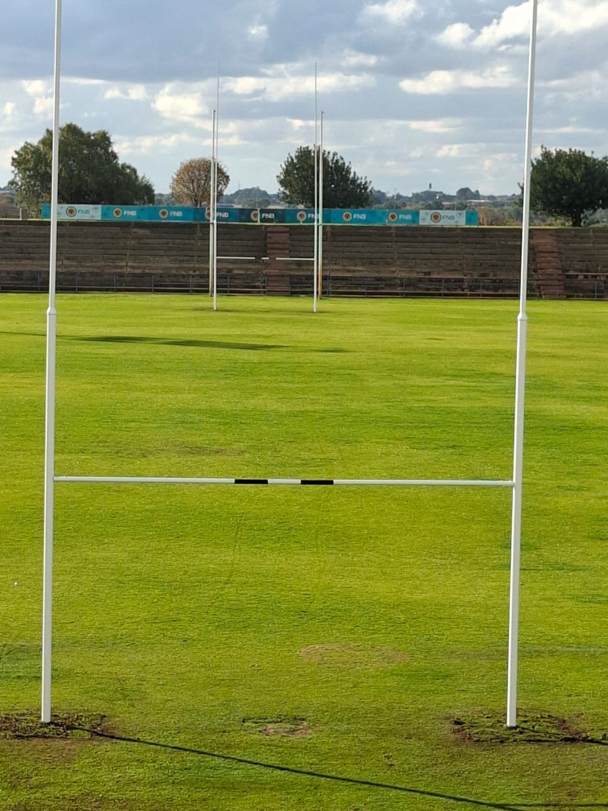 14 rugby posts installed 1 | Synsport Rugby goalposts on a bright green field with empty wooden stands and blue advertising banners behind the goalposts in the distance.