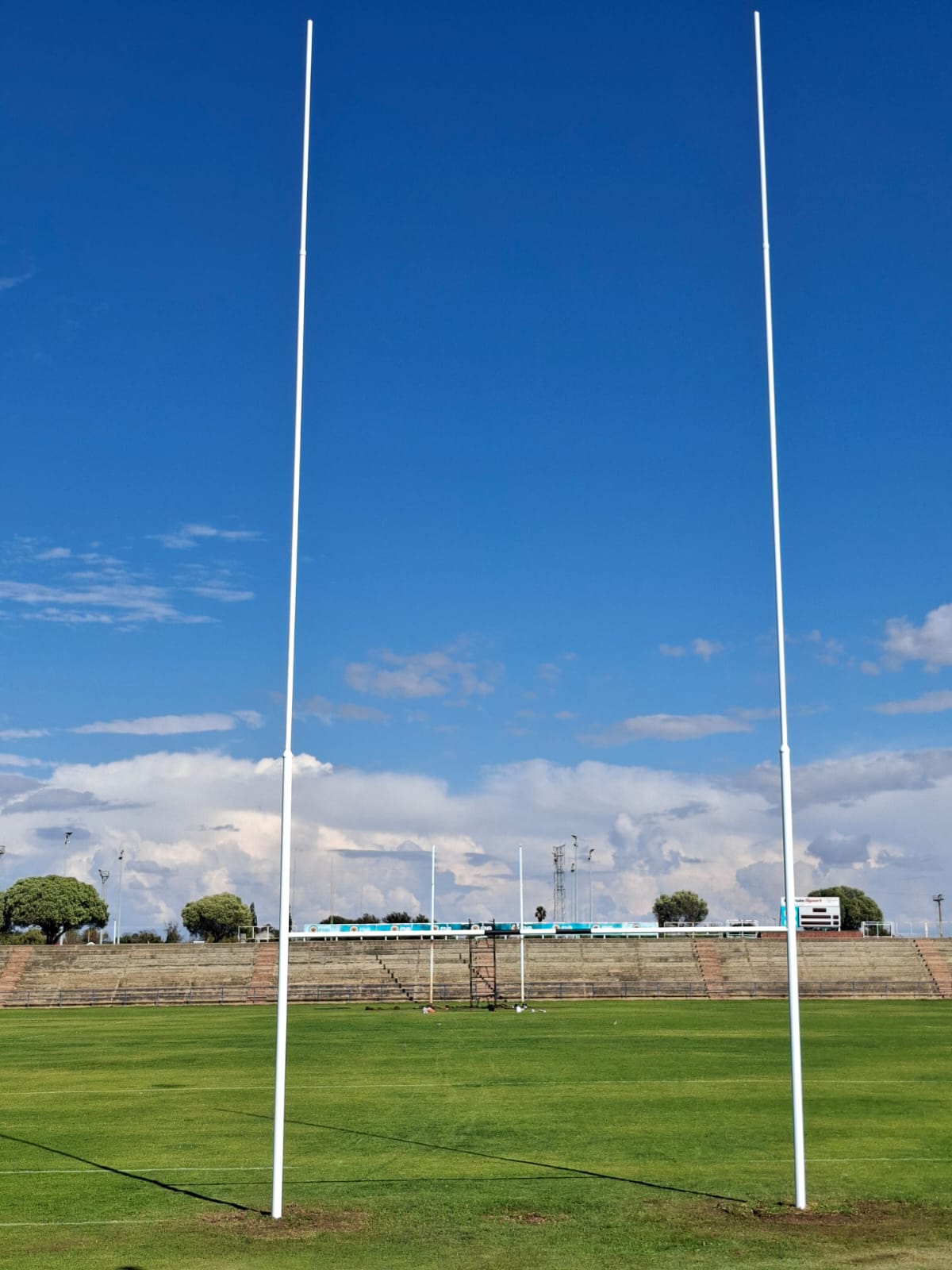 14 rugby posts installed 2 | Synsport Two tall white rugby goalposts stand on a grassy field with empty stands and a bright blue sky.