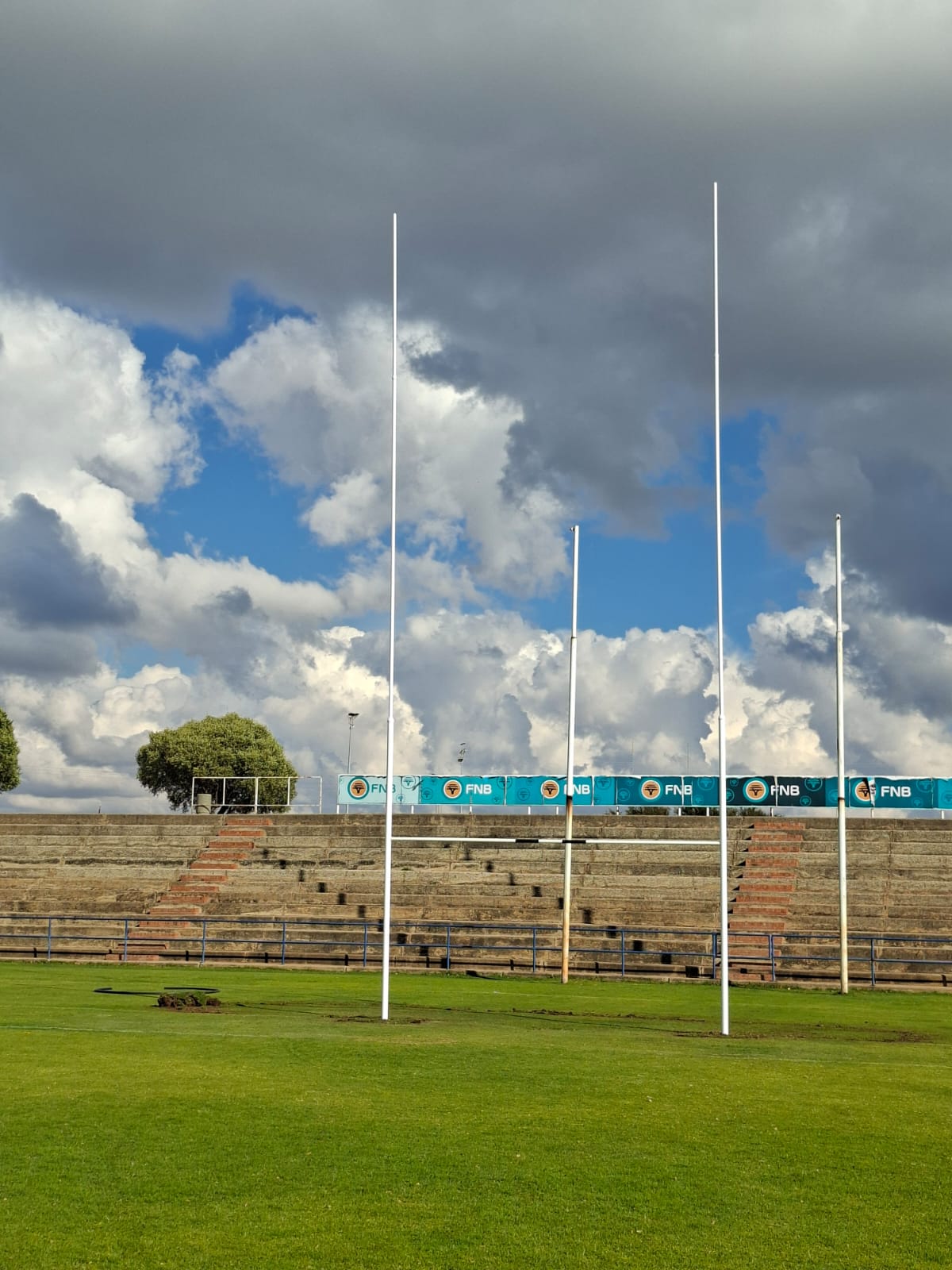 14 rugby posts installed 5 | Synsport Empty stadium field with tall white goal posts, tiered seating in the background, and blue FNB banners along the stands under a cloudy sky, bright grass.