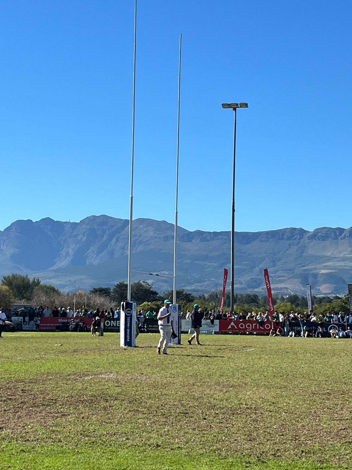 14m high rugby poles - Paarl Boys High School | Synsport Sports event on a grassy field with spectators along the sidelines and banners, mountains in the distance under a clear blue sky.