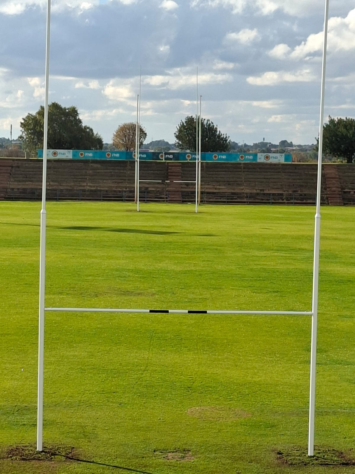 14m rugby posts installed 4 | Synsport Rugby goal posts on a green grass field with a wooden spectator stand and blue sponsor banners in the background.