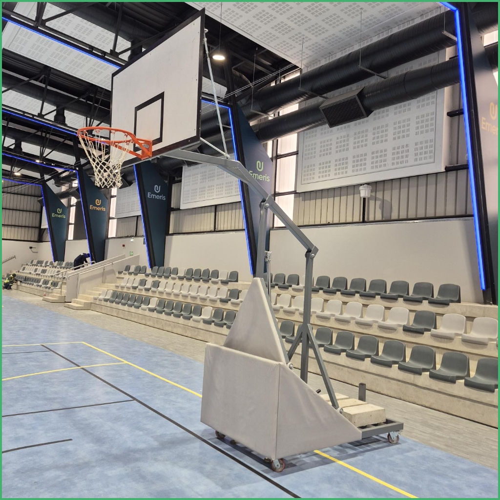 Synsport sports equipment - Basketball Folding Club 5 | Synsport Portable basketball hoop with a padded, wheeled base on a blue gym floor and empty tiered seating along the wall. The arena features banners and visible ductwork above in a well-lit indoor sports hall.