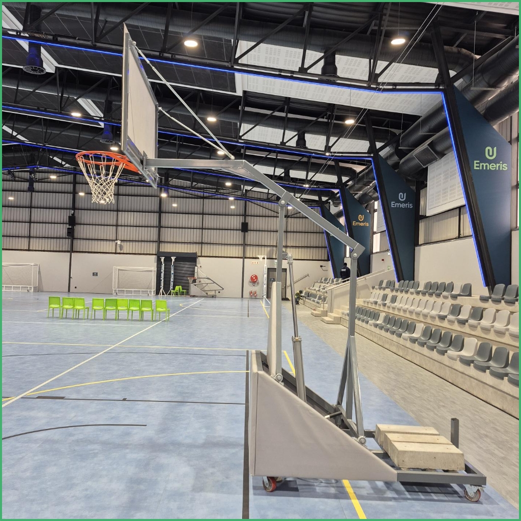 Synsport sports equipment - Basketball Folding Club 6 | Synsport Indoor sports hall with a movable basketball hoop on wheels, blue court, and gray bleachers along the right.
