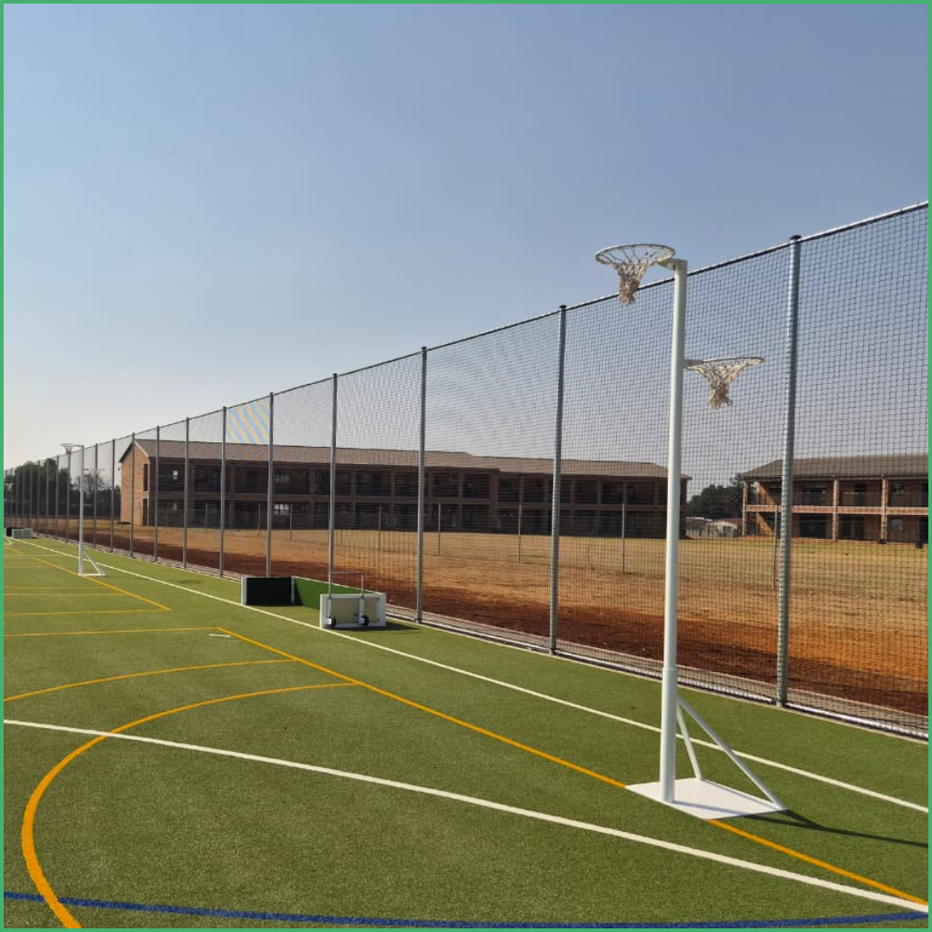 Synsport sports equipment - Netball double height Frestanding 05 | Synsport Outdoor multiuse sports court with green turf, white and yellow lines, tall chain-link fence, and two basketball hoops by the fence; school buildings in background.
