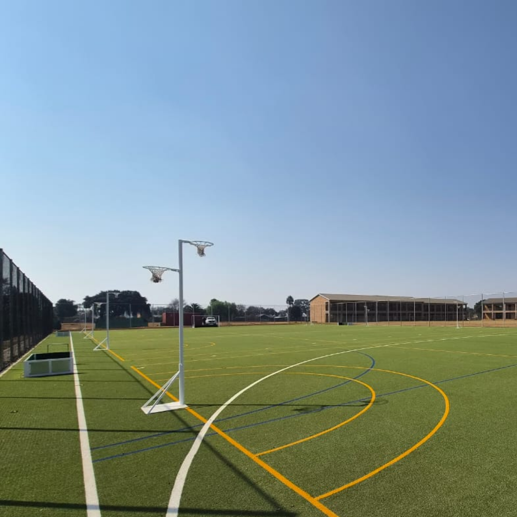 Synsport sports equipment - Netball double height Frestanding 07 | Synsport Wide view of an outdoor artificial turf field with multiple sport lines, two basketball hoops on the left, a fence, and distant school buildings under a clear blue sky.