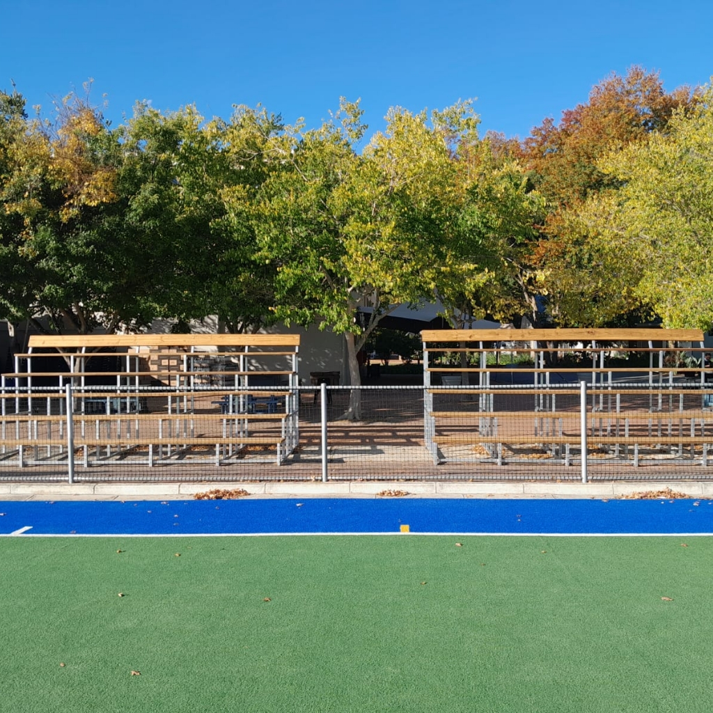 Synsport sports equipment - T-1 | Synsport Empty wooden bleachers behind a metal fence along a green turf field with a blue track and autumn trees under a clear blue sky.