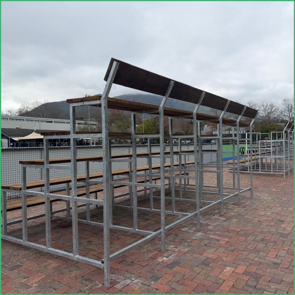 Synsport sports equipment - T-2 | Synsport Outdoor metal stadium bleachers with wooden benches on a brick-paved area beside a sports field.