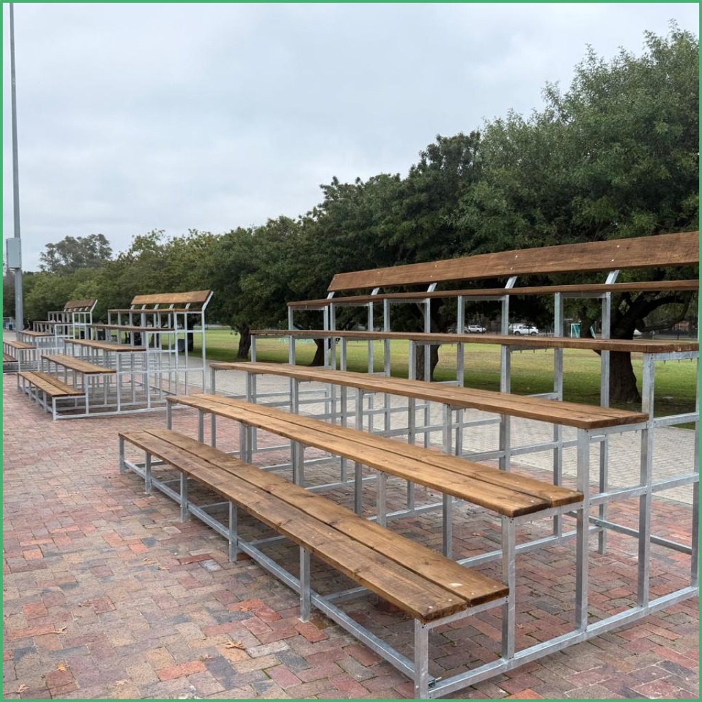 Synsport sports equipment - T-3 | Synsport Outdoor wooden bleachers with metal framing on a brick-paved area, trees in the background at a park or stadium entrance.