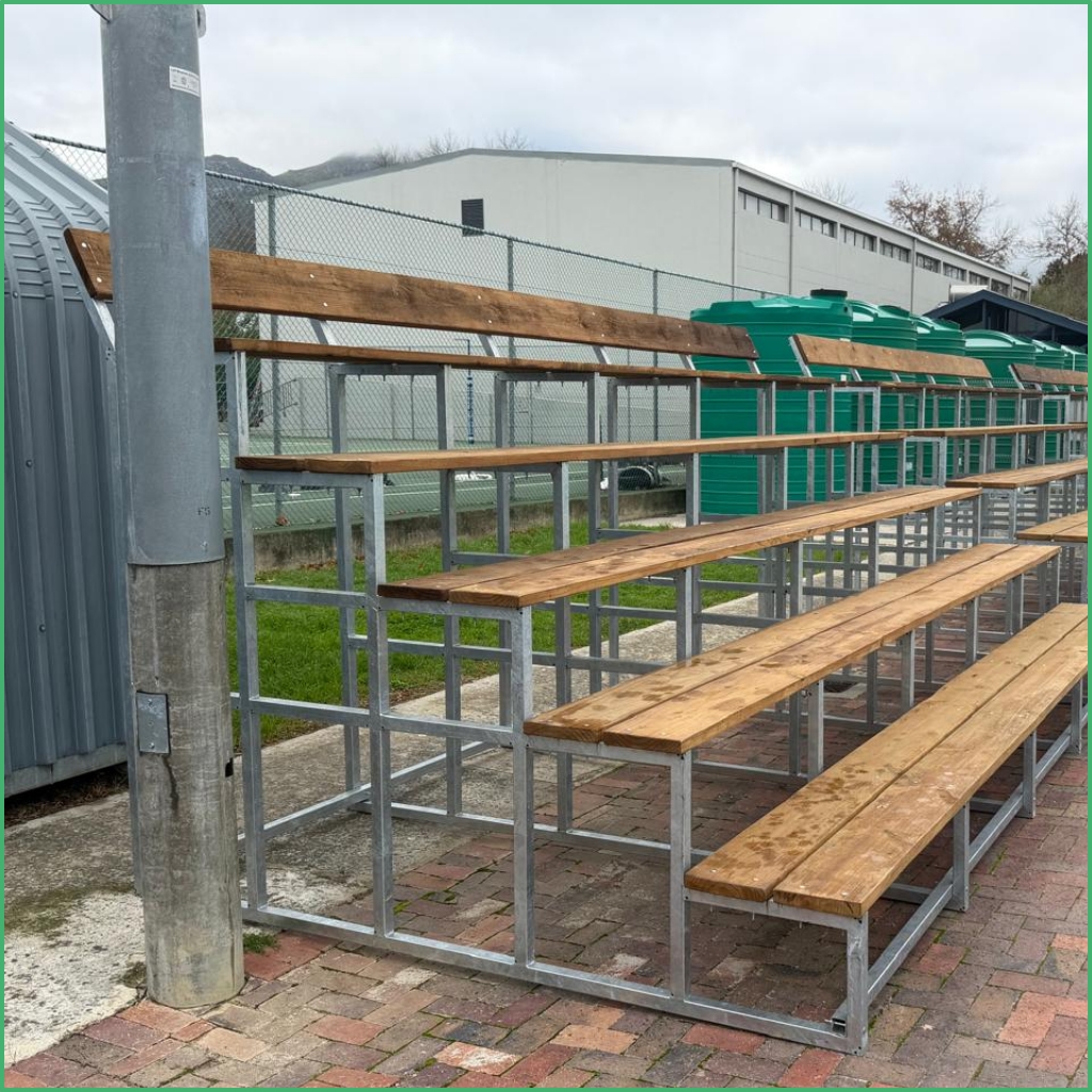 Synsport sports equipment - T-4 | Synsport Outdoor wooden bleachers with a metal frame beside a chain-link fence and green waste bins in the background, near a light-gray building.