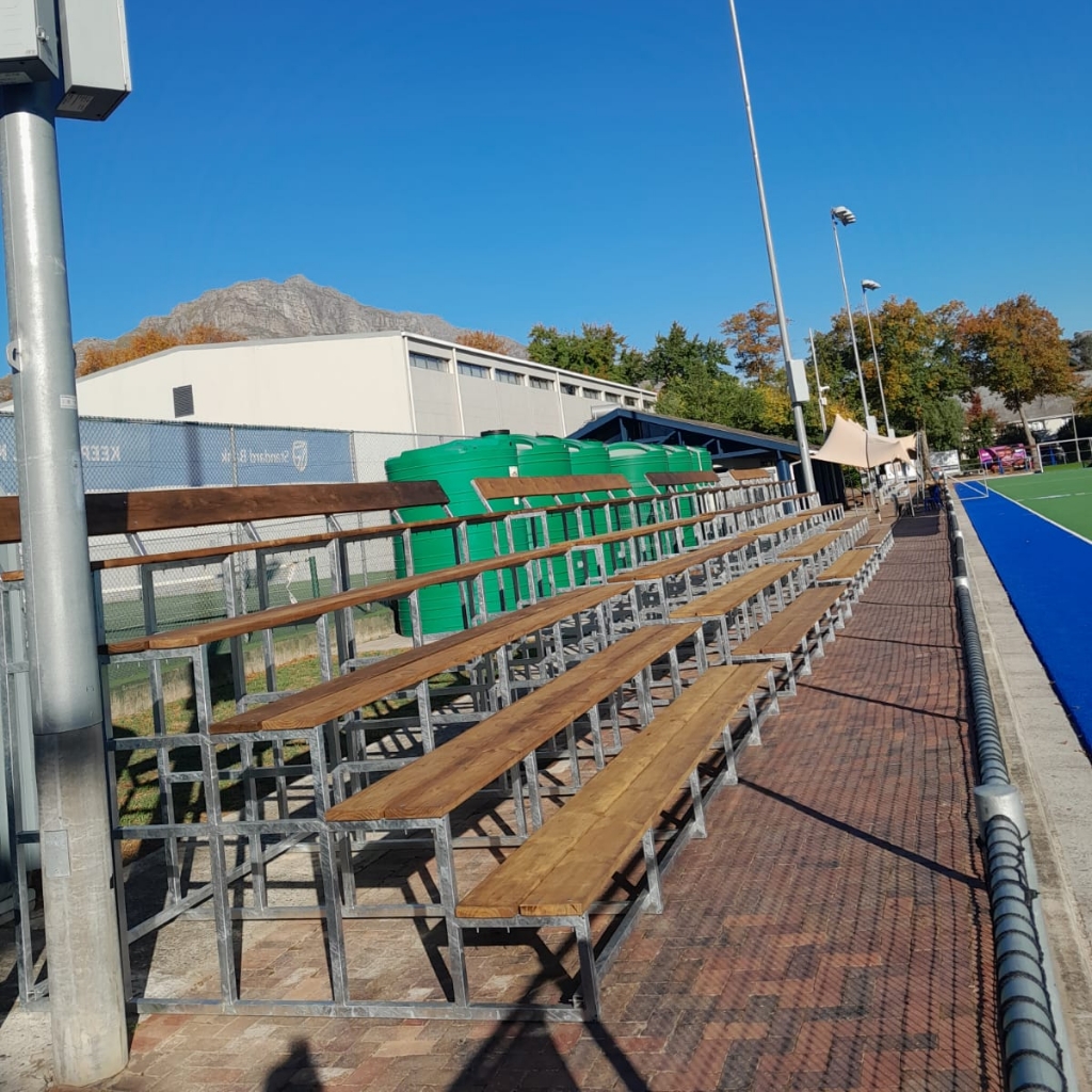 Synsport sports equipment - T | Synsport Empty stadium bleachers with wooden benches and metal frames beside a blue track on a sunny day, with green bins nearby and mountains in the background.