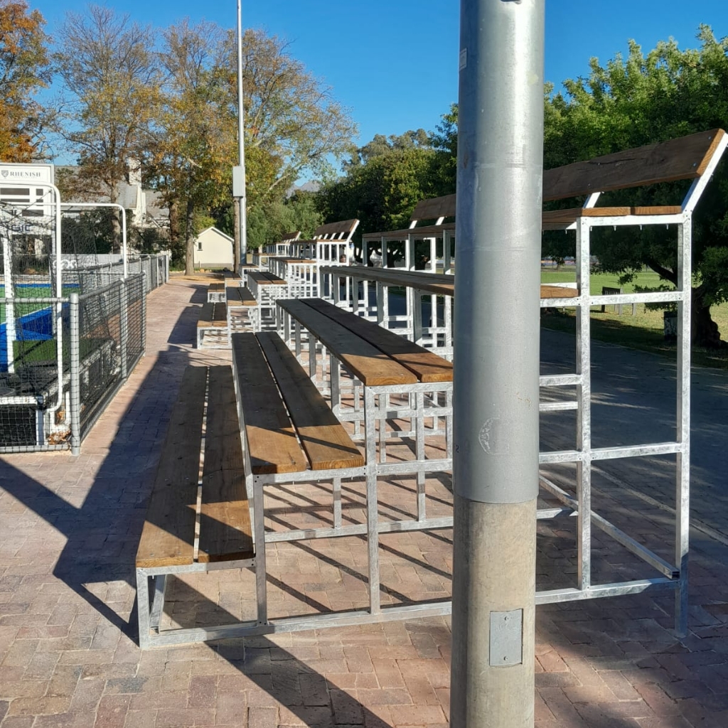 Synsport sports equipment - Ti | Synsport Outdoor metal bleachers with wooden benches along a brick path under a clear blue sky, in a sunny park area.