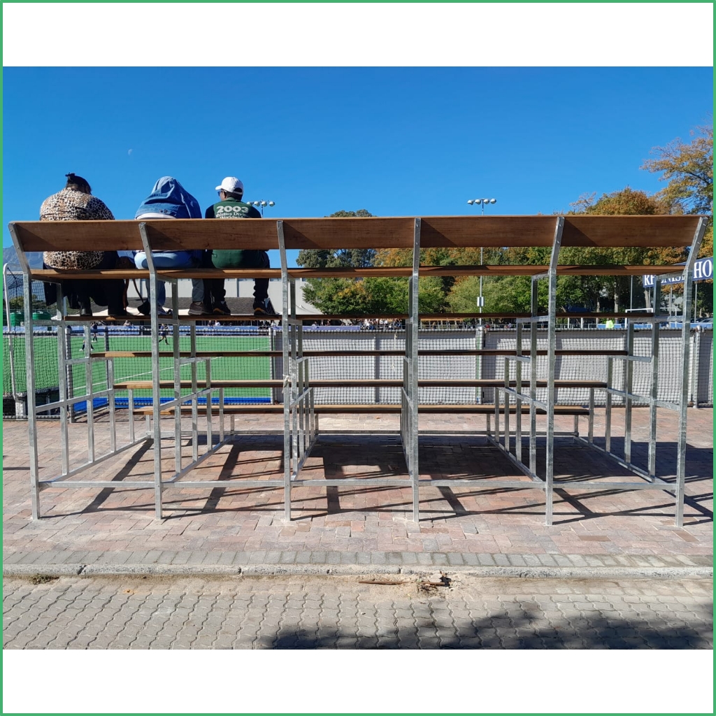 Synsport sports equipment - Tie | Synsport Three people sitting on wooden bleachers with metal frames overlooking a sunny sports field.