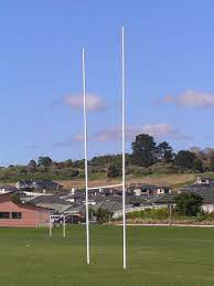 GT Rugby Posts 10m Standard Mild Steel 2 | Synsport Two tall white rugby goal posts rise on a green field, with a row of houses on a hill in the background.