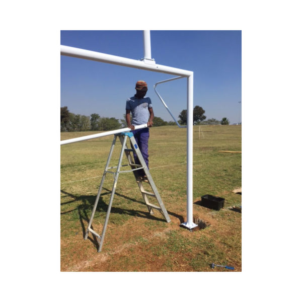 GT soccer rugby combination posts 2 | Synsport Man on a ladder assembling a large white metal goalpost frame in an open field under a clear blue sky.