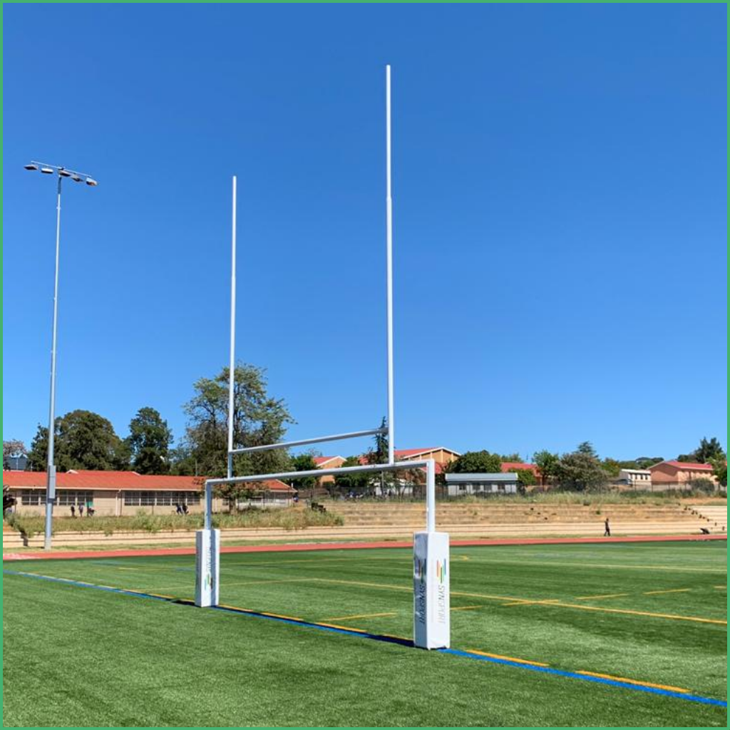 Synsport sports equipment - GT Rugby Soccer Combo | Synsport Rugby goalposts on a green turf field with school buildings and trees in the background under a clear blue sky.