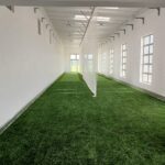 Synthetic Cricket Facilities Milnerton HS - Indoor cricket nets x 2 | Synsport Indoor cricket practice lane with netted walls and artificial turf leading to bright doorway at far end, multiple windowed walls on the right.