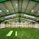 Synthetic Cricket Facilities Pearson Indoor | Synsport Indoor sports facility with artificial turf, green steel beams, and central netted practice cages under bright overhead lights.