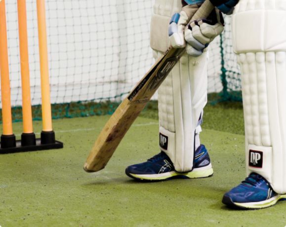 Synthetic Cricket Facilities-img2 | Synsport Cricket batter wearing pads and gloves with a bat resting on the ground beside orange stumps in a practice nets area, green artificial turf below. (informative)