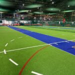 Synthetic Cricket Facilities no name 2 | Synsport Indoor sports facility with green turf, blue pitch lanes, cricket stumps on the right, and seating along the back wall at the far end.