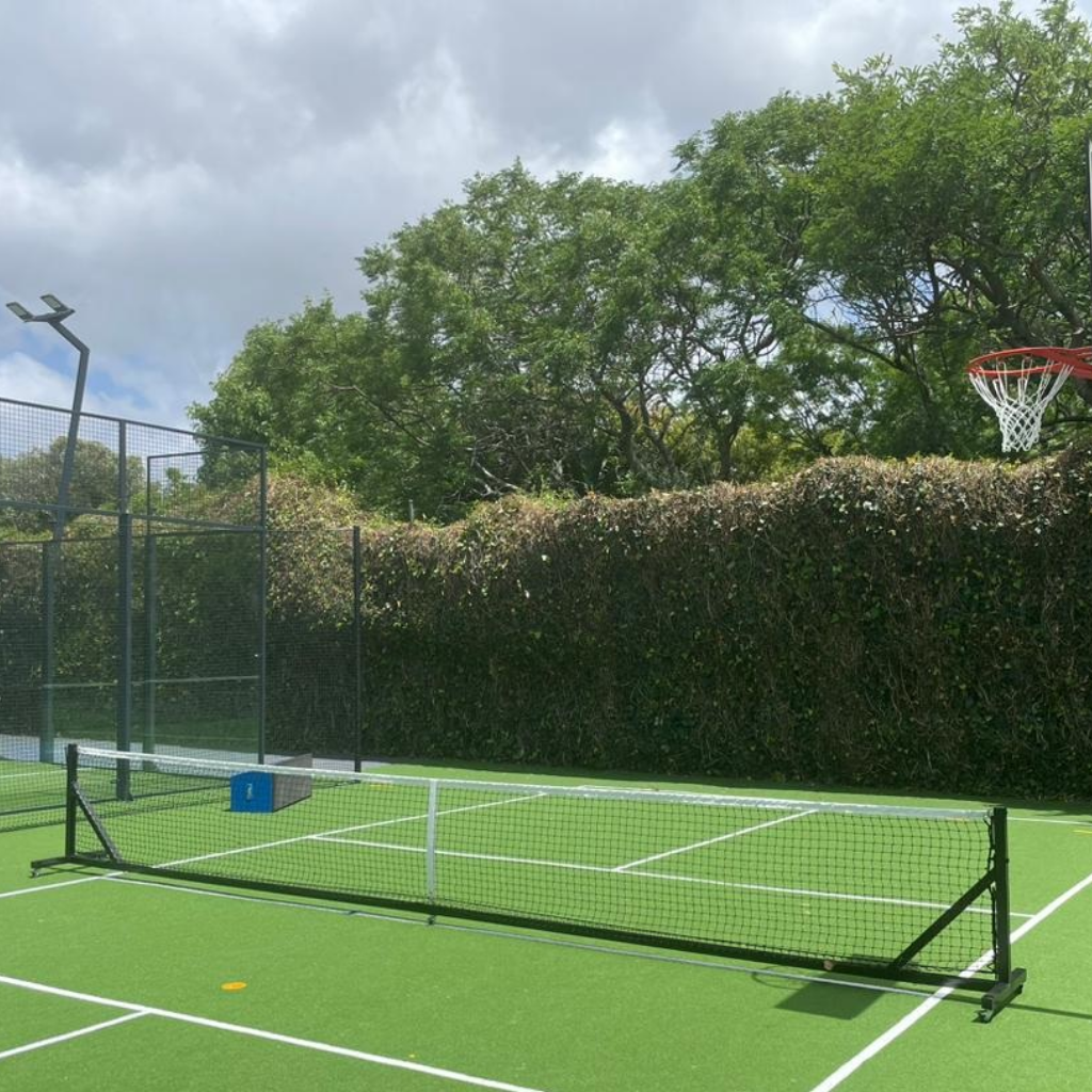 Synsport sports equipment - Free standing pickleball posts and net 2 | Synsport Outdoor tennis/mini-court with a low net, surrounded by a tall fence and dense hedge, and a basketball hoop visible to the right under a cloudy sky.