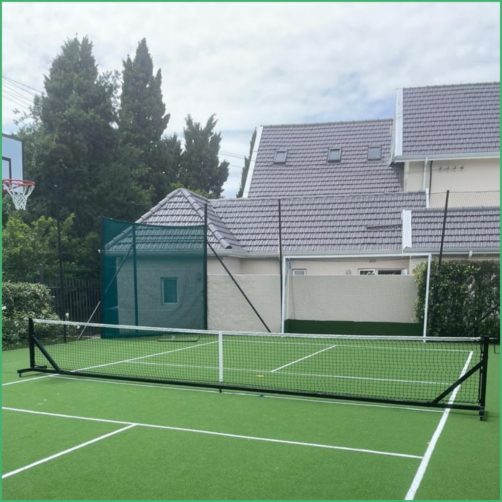 Synsport sports equipment - Free standing pickleball posts and net | Synsport Outdoor tennis court with a net on a green surface, white boundary lines, and a basketball hoop visible to the left behind a fence.